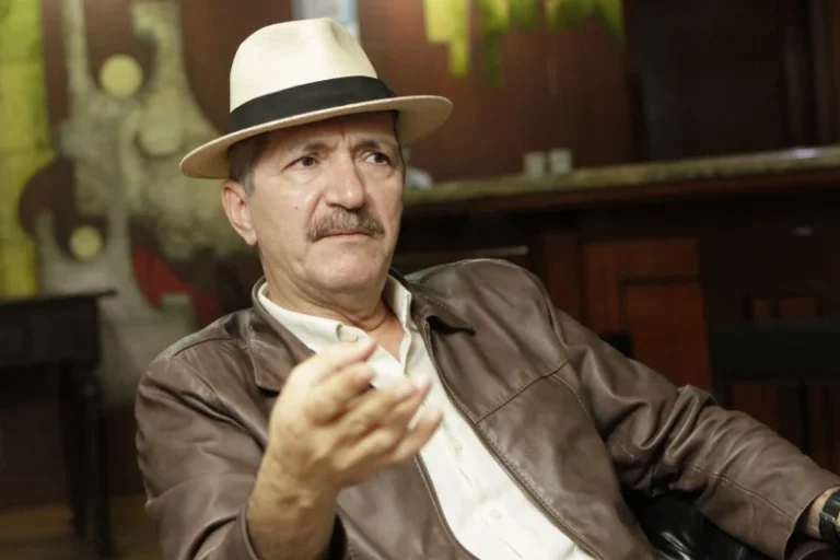 Aldo Rebelo sentado, usando chapéu claro com faixa preta, jaqueta de couro marrom e camisa clara, com uma das mãos erguida.