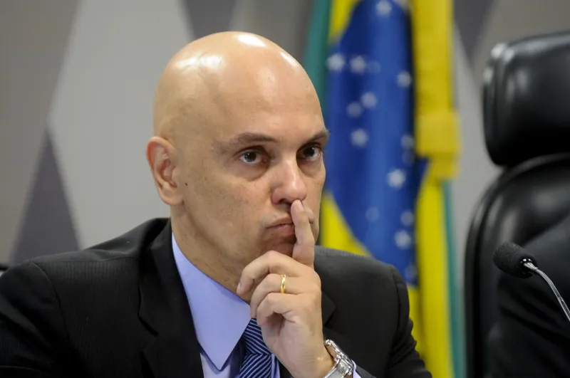 Alexandre de Moraes, de terno escuro e gravata azul, mantém o dedo indicador próximo aos lábios, com a bandeira do Brasil ao fundo.