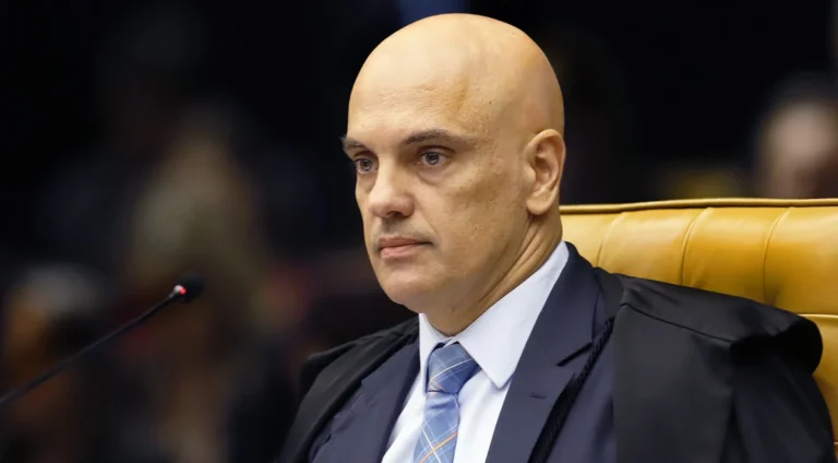 Homem de cabeça raspada, usando toga preta sobre terno escuro e gravata azul, sentado diante de um microfone, com cadeira amarela ao fundo.