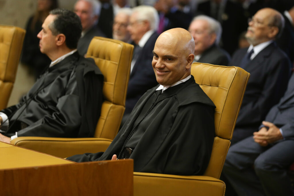 Alexandre de Moraes sorri sentado em uma cadeira amarela, usando toga preta, durante cerimônia no STF, com outros magistrados ao fundo.