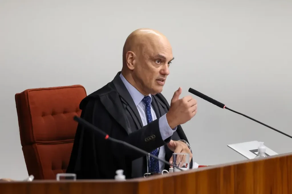 Alexandre de Moraes, de toga preta, camisa clara e gravata azul, está sentado em uma cadeira vermelha durante sessão do STF, falando ao microfone e gesticulando com a mão direita.