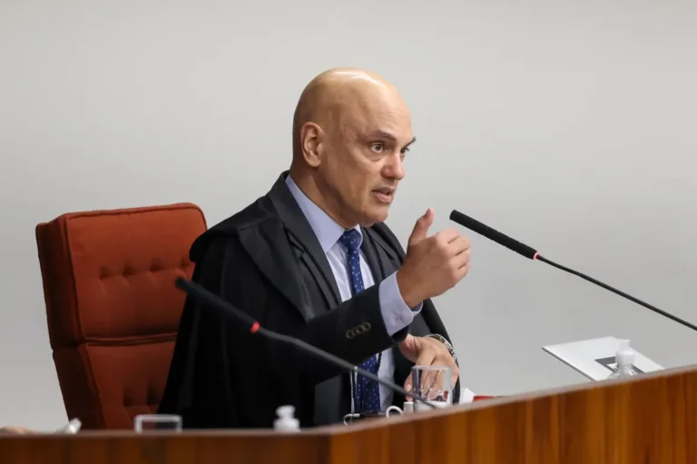 Alexandre de Moraes, de toga preta, camisa clara e gravata azul, está sentado em uma cadeira vermelha durante sessão do STF, falando ao microfone e gesticulando com a mão direita.