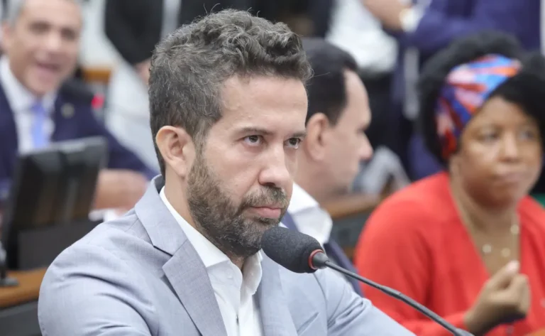 André Janones, de paletó cinza-claro e camisa branca, diante de um microfone, com outras pessoas ao fundo.