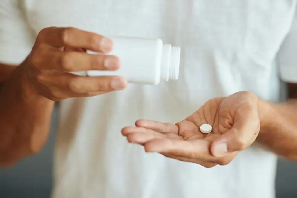 Pessoa de camiseta branca segura um frasco de medicamento em uma mão e exibe um comprimido branco na palma da outra.