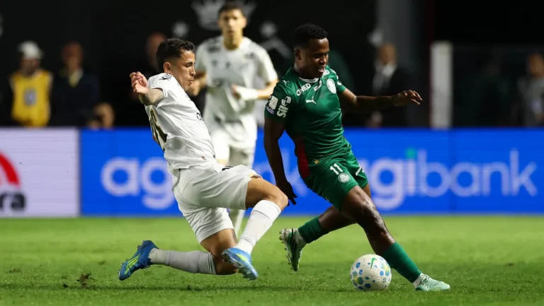 Arias, de uniforme verde do Palmeiras, conduz a bola enquanto jogador do Santos, de branco, tenta o desarme com um carrinho.