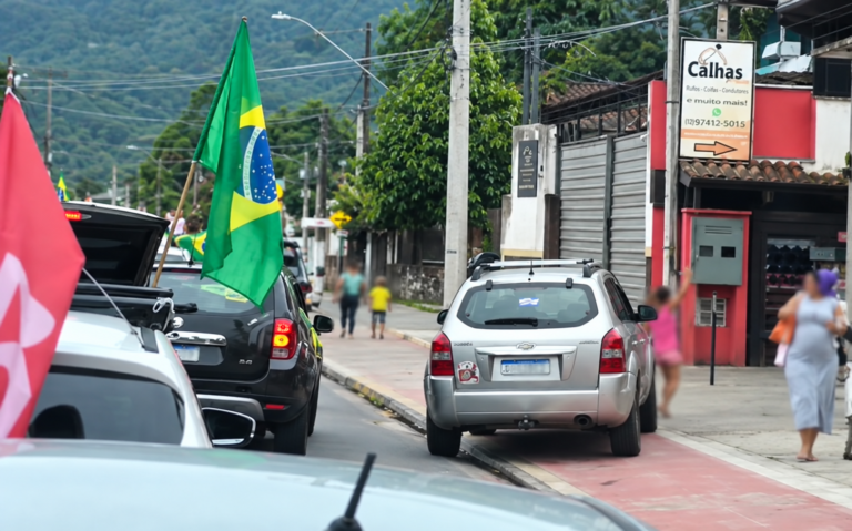 Carro prateado junto à calçada, bandeira do Brasil em outro veículo e criança desfocada ao lado do automóvel.