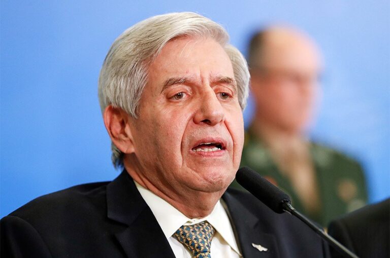 Augusto Heleno, de terno escuro e gravata estampada, fala diante de um microfone, com uma pessoa desfocada ao fundo.