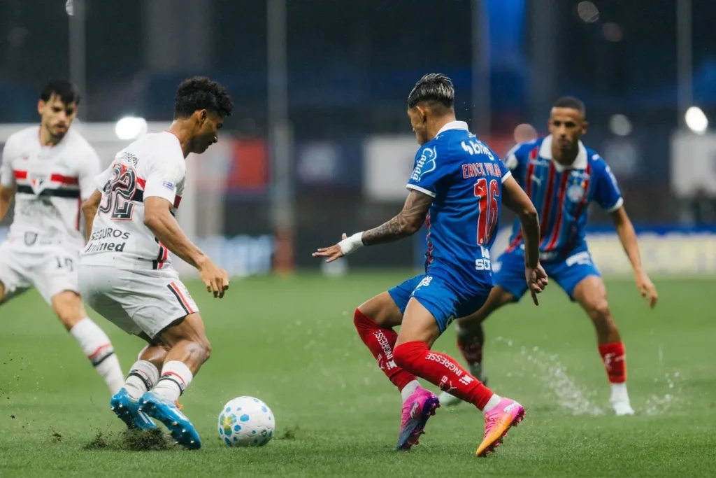 Jogador do São Paulo, de uniforme branco, disputa a bola com atleta do Bahia, de azul e vermelho, sobre o gramado molhado, com outros jogadores ao fundo.