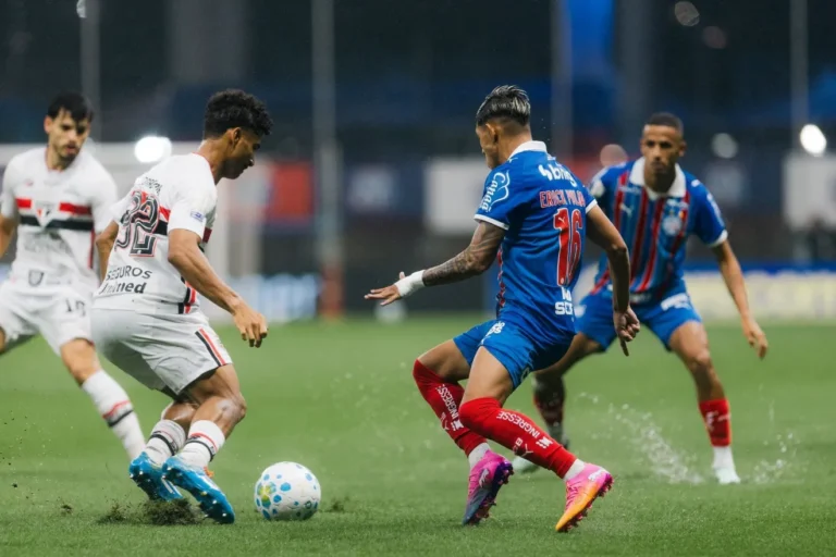 Jogador do São Paulo, de uniforme branco, disputa a bola com atleta do Bahia, de azul e vermelho, sobre o gramado molhado, com outros jogadores ao fundo.