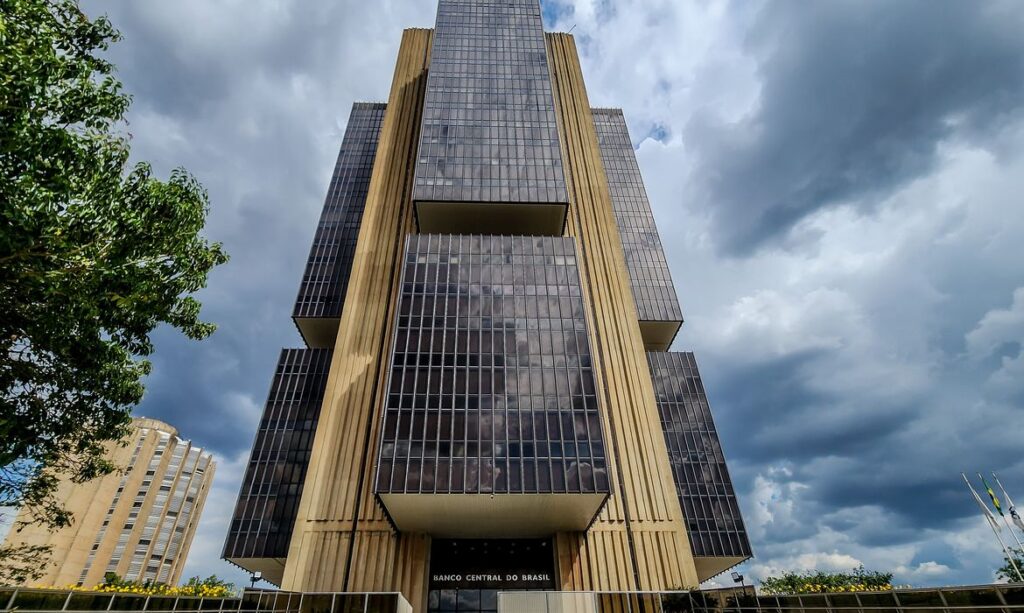 Sede do Banco Central do Brasil, em Brasília, vista de baixo, com fachada envidraçada, colunas verticais e céu nublado ao fundo.