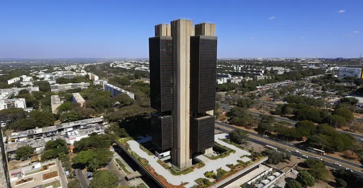 Vista aérea da sede do Banco Central em Brasília, com torres de fachadas escuras, cercada por ruas, árvores e outros edifícios.