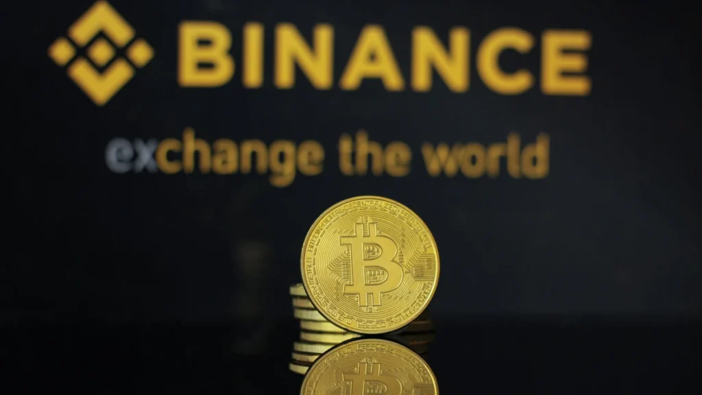 Moeda dourada de Bitcoin sobre uma superfície escura, com o logotipo amarelo da Binance desfocado ao fundo.