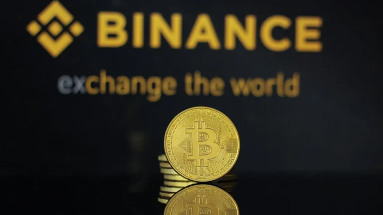 Moeda dourada de Bitcoin sobre uma superfície escura, com o logotipo amarelo da Binance desfocado ao fundo.