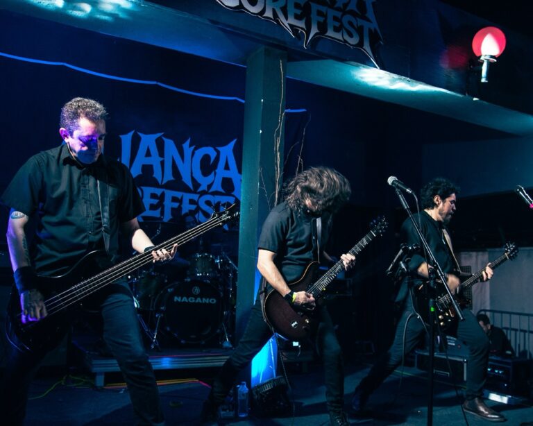 Três músicos vestidos de preto tocam baixo e guitarras em um palco iluminado por luzes azuis, com bateria ao fundo.
