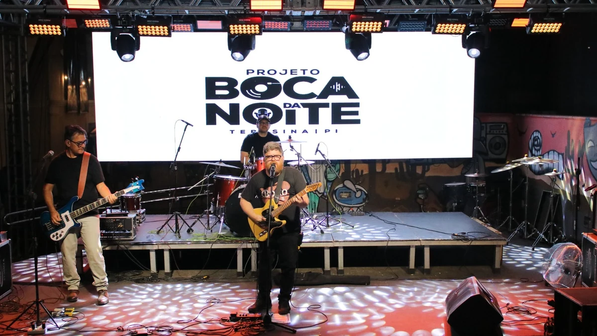 Três músicos se apresentam em um palco com guitarras, baixo e bateria, diante de um painel do projeto Boca da Noite, em Teresina.