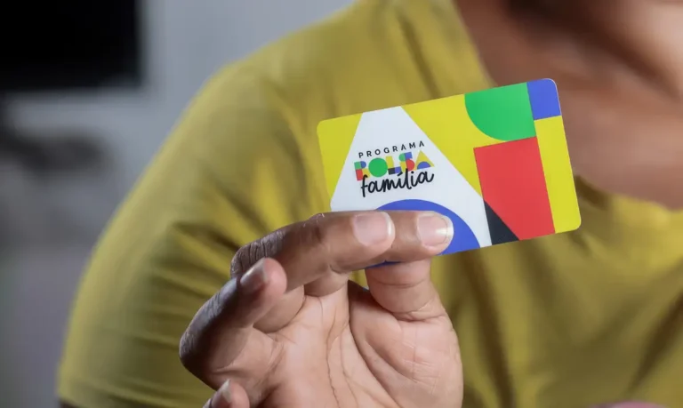 Mão segurando cartão colorido do programa Bolsa Família, diante de uma pessoa de camiseta amarela.
