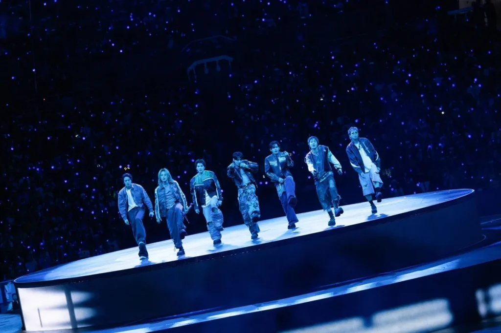 Sete integrantes do BTS dançam sobre uma passarela iluminada durante show, diante de uma multidão com luzes azuis na plateia.