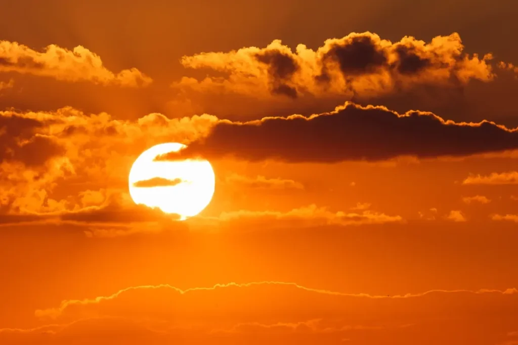 Sol brilhante próximo ao horizonte, parcialmente encoberto por nuvens escuras, em um céu de tons alaranjados.
