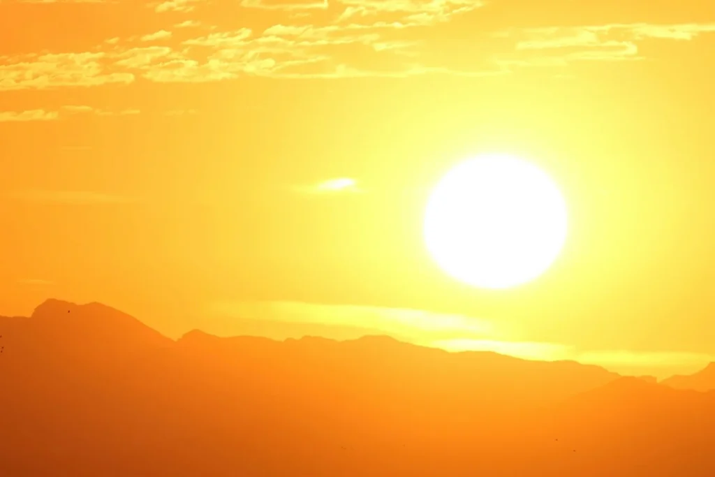 Sol intenso em céu amarelo e alaranjado, iluminando uma paisagem de serras no horizonte.