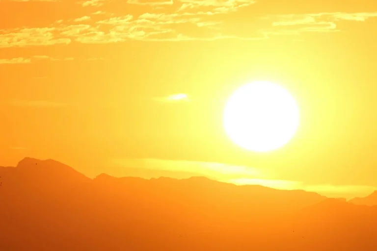 Sol intenso em céu amarelo e alaranjado, iluminando uma paisagem de serras no horizonte.