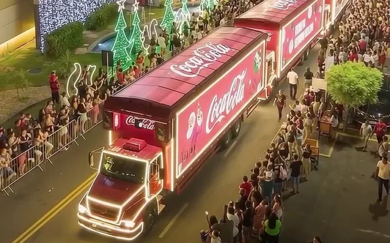 Caminhões vermelhos iluminados da Coca-Cola, com pessoas nas laterais da rua e decoração natalina.