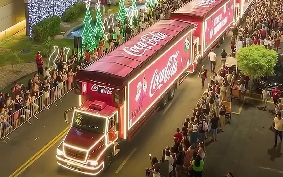 Caminhões vermelhos iluminados da Coca-Cola, com pessoas nas laterais da rua e decoração natalina.