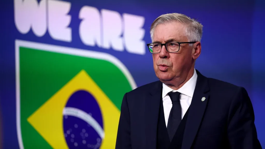 Carlo Ancelotti, de terno escuro e óculos, diante de um painel azul com a bandeira do Brasil ao fundo.