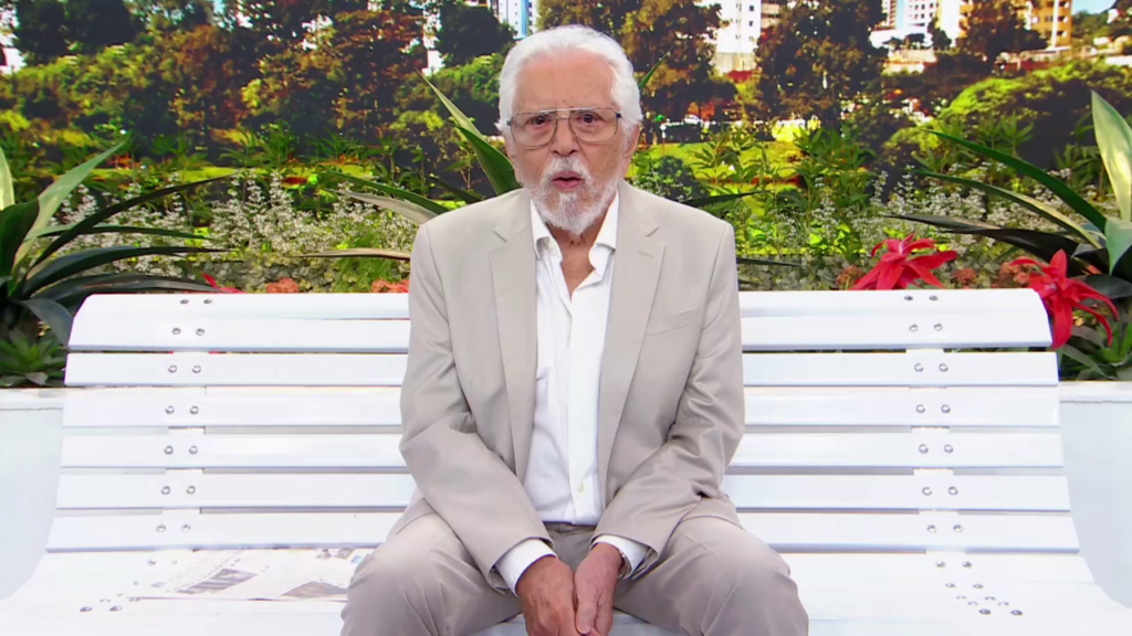 Carlos Alberto de Nóbrega, de terno bege e camisa branca, sentado em um banco diante de um jardim, com prédios ao fundo.