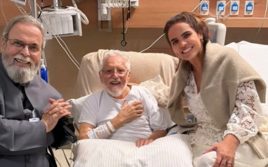 Carlos Alberto de Nóbrega sentado em leito hospitalar, ao lado do padre Antônio Maria e de Renata de Nóbrega.