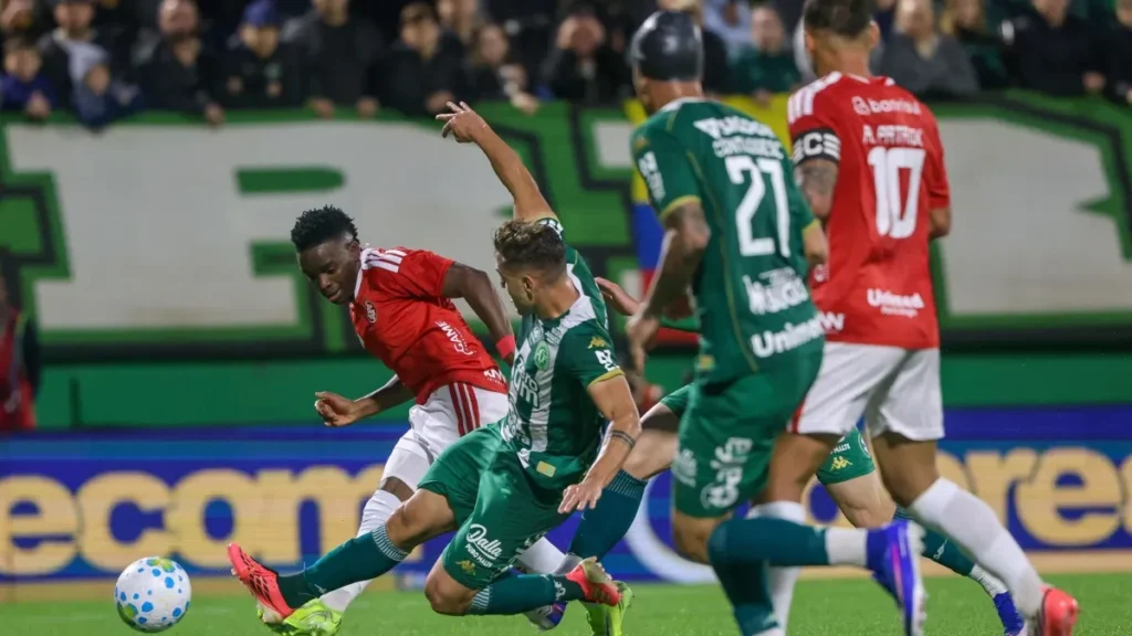 Jogador do Internacional, de uniforme vermelho, disputa a bola com atleta da Chapecoense, de verde, que estica a perna para tentar o desarme, enquanto outros jogadores acompanham o lance.