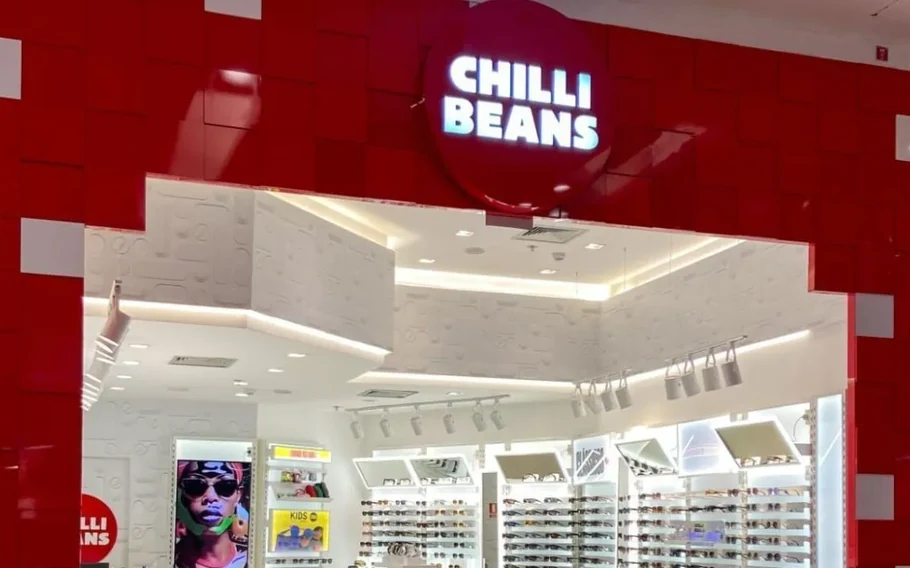 Fachada vermelha de uma loja Chilli Beans, com letreiro luminoso e prateleiras de óculos visíveis no interior.
