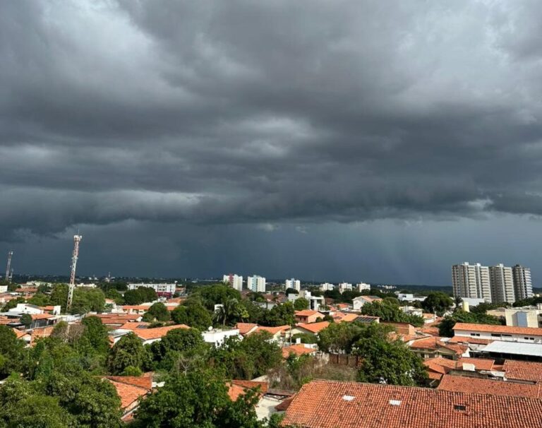 Nuvens escuras cobrem área urbana com casas, árvores e prédios ao fundo, indicando tempo instável.