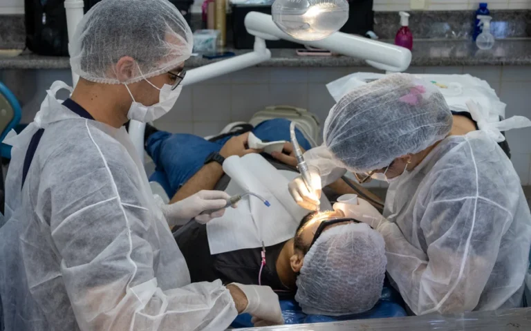 Dois profissionais com toucas, máscaras e aventais atendem paciente em cadeira odontológica.