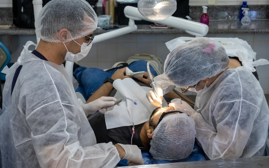 Dois profissionais com toucas, máscaras e aventais atendem paciente em cadeira odontológica.