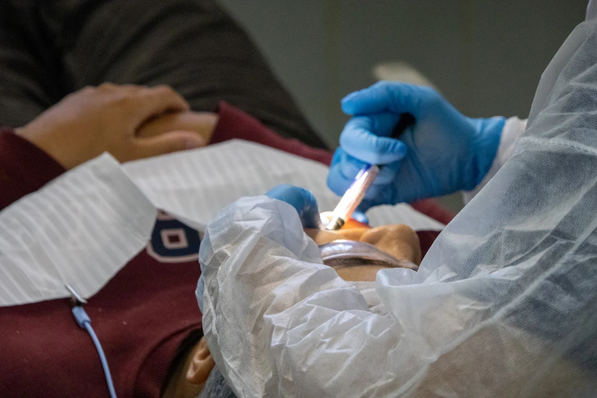 Profissional com luvas azuis utiliza instrumento odontológico iluminado na boca de paciente reclinado.