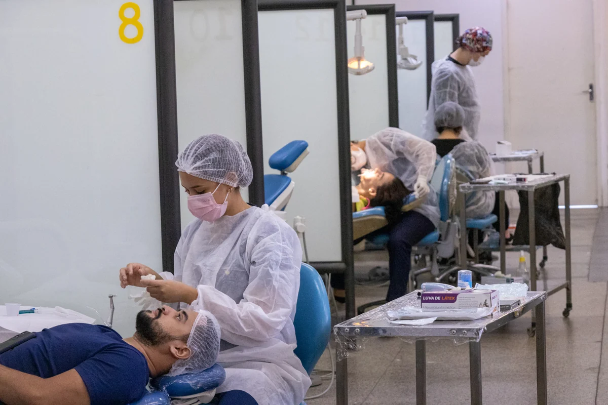 Clínica de odontologia com pacientes em cadeiras de atendimento, profissionais com máscaras e toucas e divisórias entre os espaços.