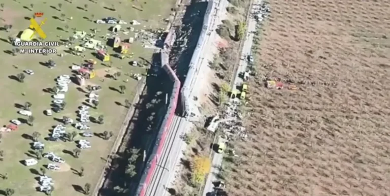 Vista aérea de um trem junto aos trilhos, com veículos de emergência e várias pessoas espalhadas por uma área aberta, ao lado de um campo agrícola na Espanha.