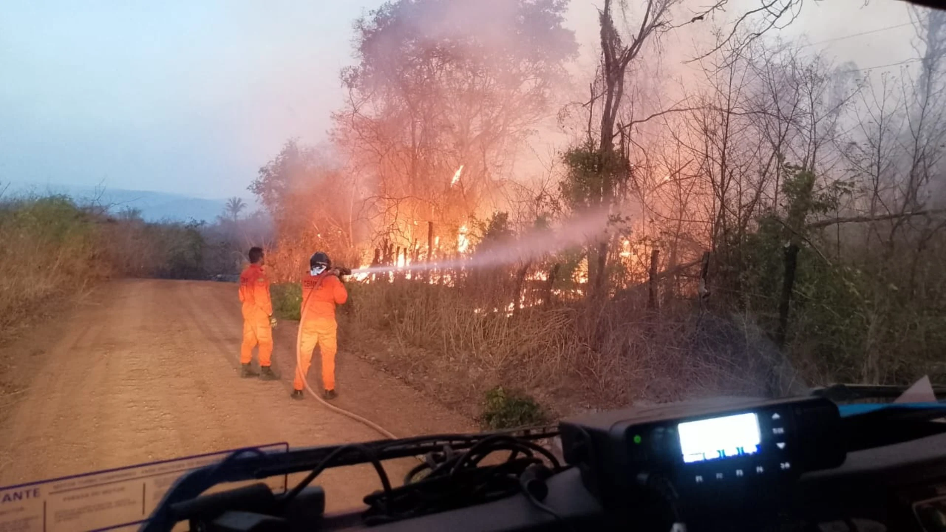 Dois agentes vestidos de laranja combatem incêndio na vegetação, à margem de uma estrada de terra.