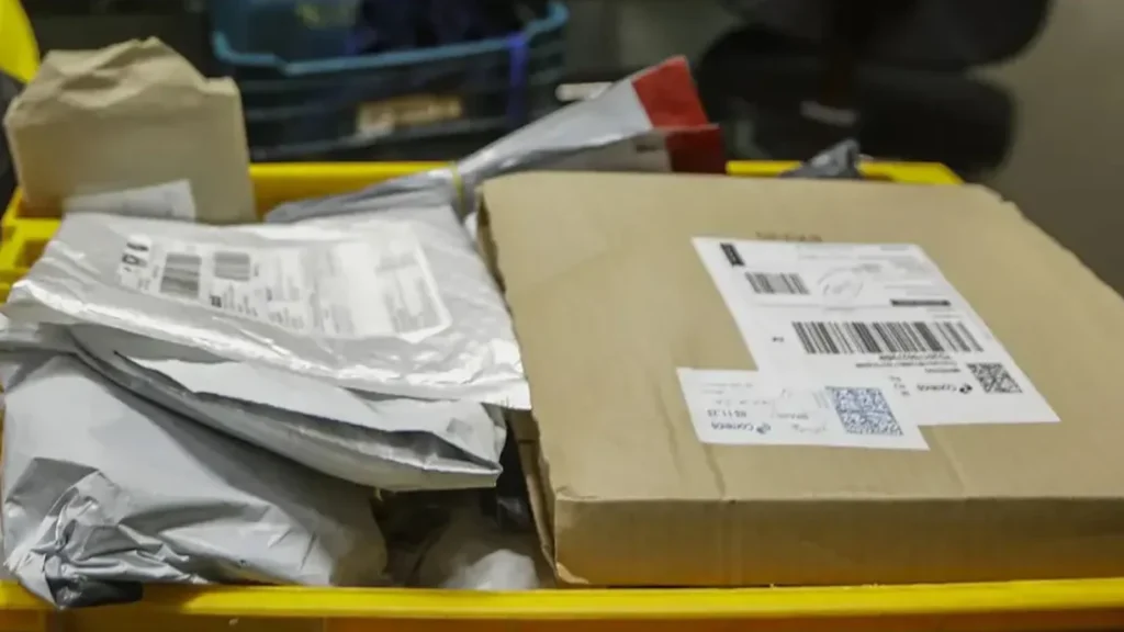 Caixa de papelão e envelopes de encomendas com etiquetas de envio e códigos de barras sobre um recipiente plástico amarelo.