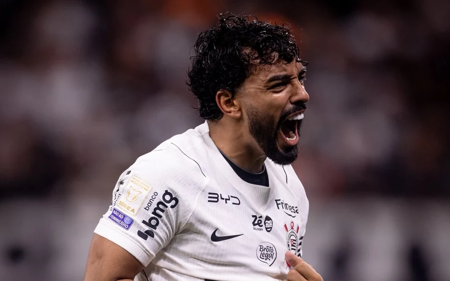 Jogador do Corinthians, de camisa branca, grita durante comemoração, com o estádio desfocado ao fundo.