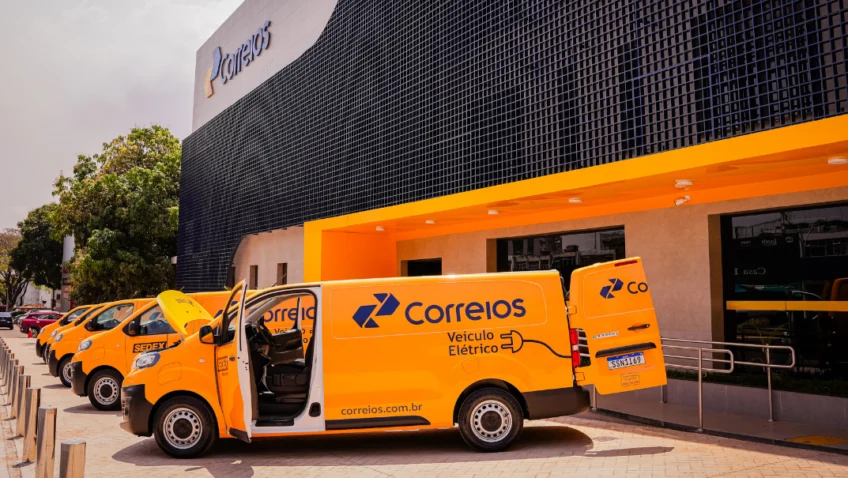 Fachada de uma unidade dos Correios com vários veículos amarelos de entrega estacionados em frente, incluindo uma van elétrica com as portas abertas.