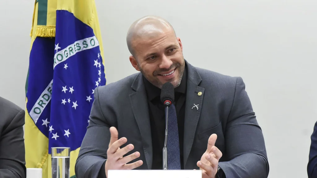 Daniel Silveira sorri e gesticula diante de um microfone, usando terno escuro, com a bandeira do Brasil ao fundo.