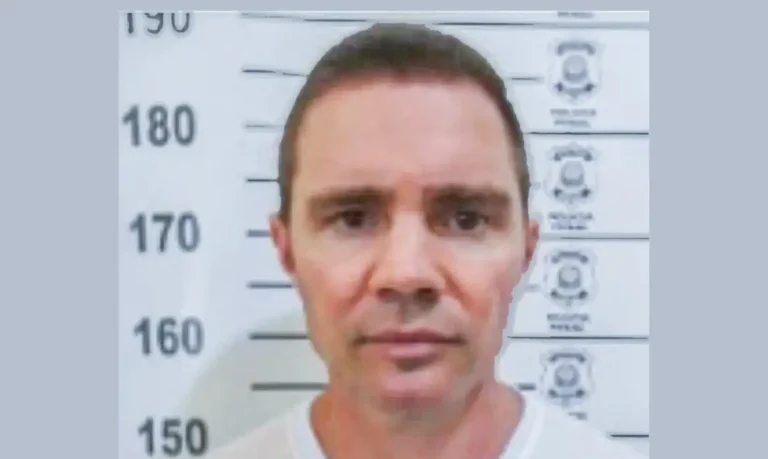 Homem de camiseta branca fotografado de frente diante de painel de identificação com marcações de altura.