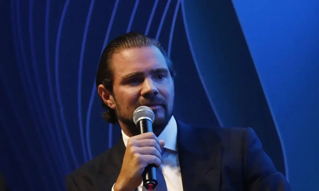 Daniel Vorcaro, de terno escuro e gravata azul, segura um microfone durante evento, diante de um fundo azul-escuro.