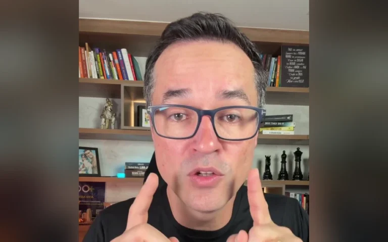Deltan Dallagnol, de óculos e camiseta preta, com os dedos indicadores erguidos diante de uma estante de livros.