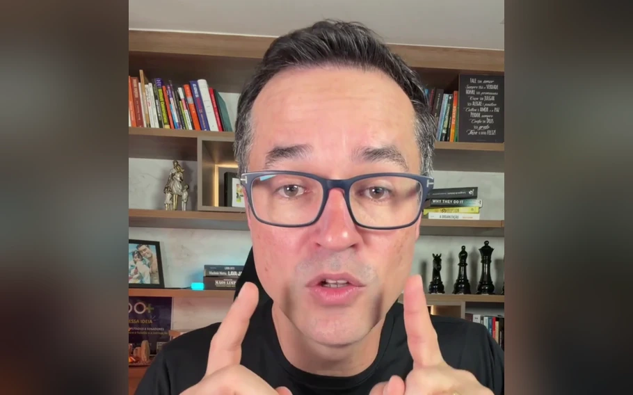 Deltan Dallagnol, de óculos e camiseta preta, com os dedos indicadores erguidos diante de uma estante de livros.