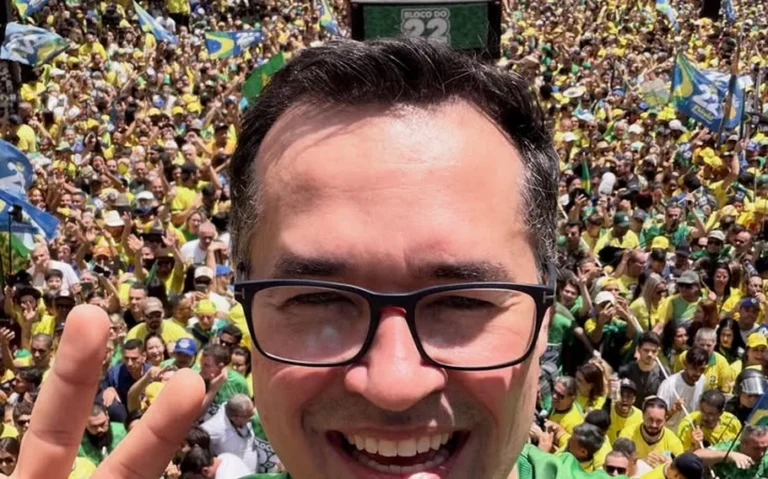 Homem de óculos e camisa verde sorri para uma selfie diante de multidão com bandeiras brasileiras.