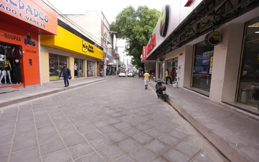 Rua comercial do Centro de Teresina, com lojas dos dois lados, pedestres, veículos ao fundo e uma motocicleta estacionada na calçada.