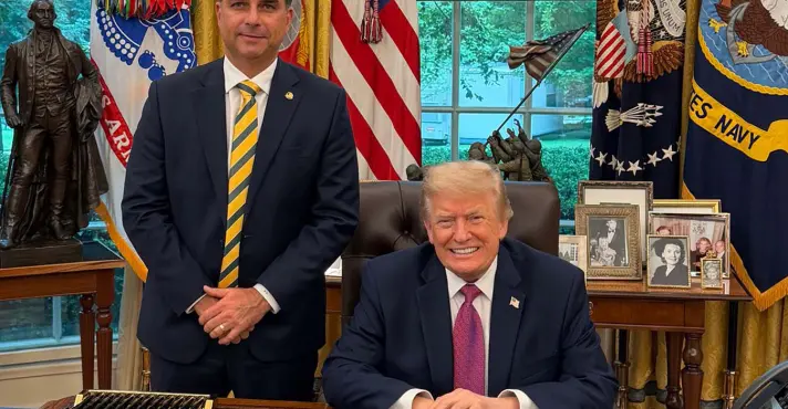 Donald Trump aparece sentado e sorridente atrás de uma mesa na Casa Branca, enquanto Flávio Bolsonaro está de pé ao seu lado, de terno azul e gravata listrada, com bandeiras americanas ao fundo.