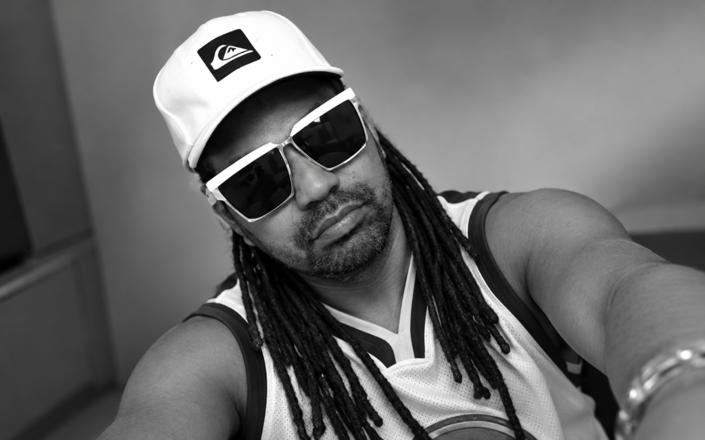 Músico e produtor cultural Edu Bizu usando boné branco, óculos escuros e cabelos em dreadlocks, em fotografia preto e branco.